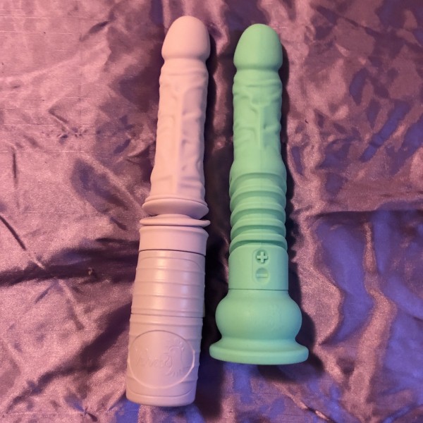 Thruster Jackie and Teddy dildos side by side. Jackie is pale purple and longer than pale green Teddy. They're on background of purple satin.